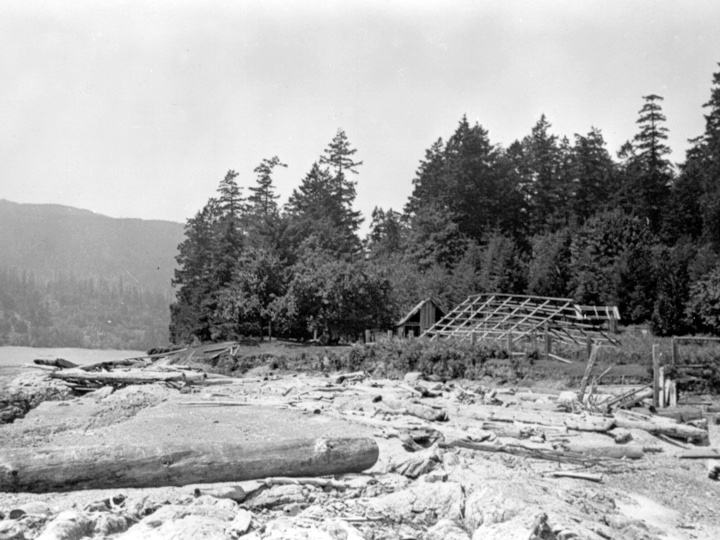 Hwu’ne’nuts in the late 20’s early 30’s, showing the ruins of Charlie Sulsulth’s gable roofed house with the longhouse in the foreground.. Salt Spring Island Archives.